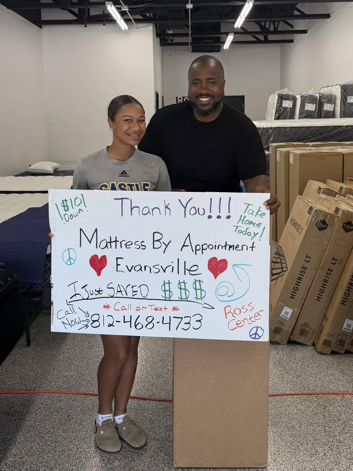 Two customers in the showroom holding a handwritten Mattress By Appointment thank-you sign