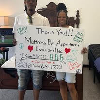 Two customers holding a handwritten thank-you sign beside a bed