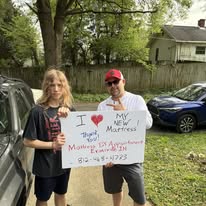 Two people outdoors holding a handwritten sign about their new mattress